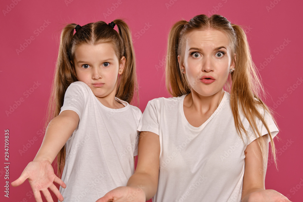 Mom and daughter with a funny ponytails, dressed in white t-shirts are posing against a pink studio background. Close-up shot. Mom and daughter with a funny ponytails, dressed in white t-shirts are posing against a pink studio background. Close-up shot.