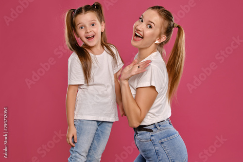 Mom and daughter with a funny ponytails, dressed in white t-shirts and blue denim jeans are posing against a pink studio background. Close-up shot.