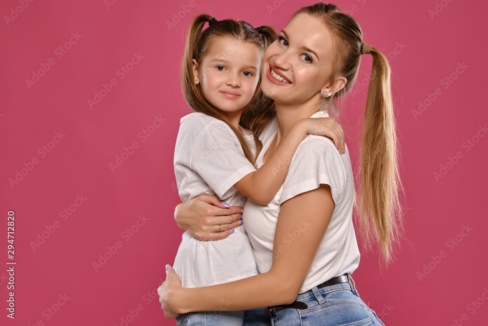 Mom and daughter with a funny ponytails, dressed in white t-shirts and blue denim jeans are posing against a pink studio background. Close-up shot. Mom and daughter with a funny ponytails, dressed in white t-shirts and blue denim jeans are posing against a pink studio background. Close-up shot.