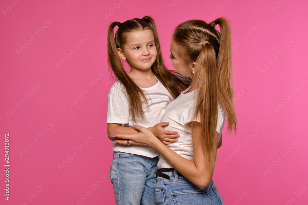 Mom and daughter with a funny ponytails, dressed in white t-shirts and blue denim jeans are posing against a pink studio background. Close-up shot. Mom and daughter with a funny ponytails, dressed in white t-shirts and blue denim jeans are posing against a pink studio background. Close-up shot.