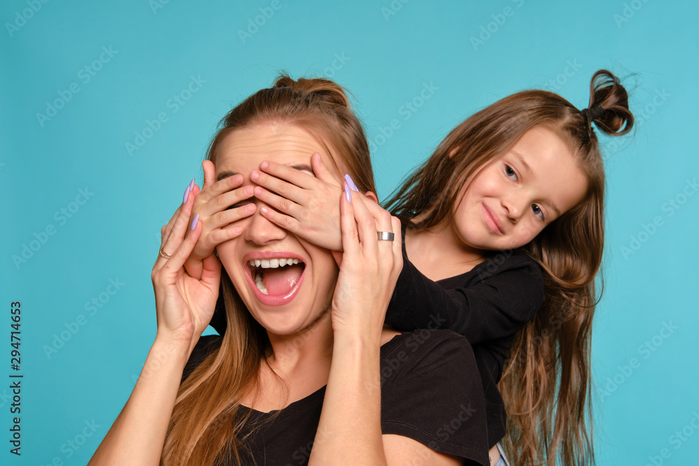 Mom and daughter with a funny hairstyles, dressed in black shirts and blue denim jeans are posing against a blue studio background. Close-up shot. Mom and daughter with a funny hairstyles, dressed in black shirts and blue denim jeans are posing against a blue studio background. Close-up shot.
