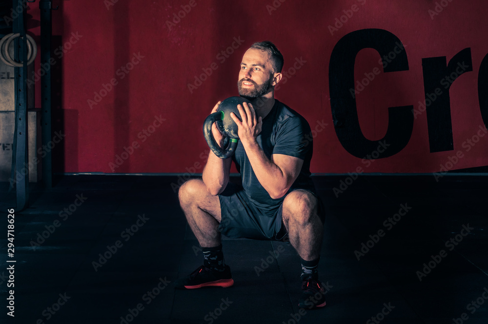 Young bearded atletic man holding kettlebell and doing squats in the modern gym. Man during workout gym. Squat with kettlebell. Sport lifestyle concept. Young bearded atletic man holding kettlebell and doing squats in the modern gym. Man during workout gym. Squat with kettlebell. Sport lifestyle concept.