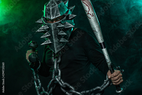 Mysterious man in a helmet with a bat and chains in black wear. Fantasy book or computer game cover concept on halloween disco party.