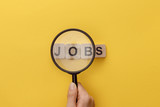 cropped view of woman holding magnifying glass under cardboard squares with jobs lettering on yellow background