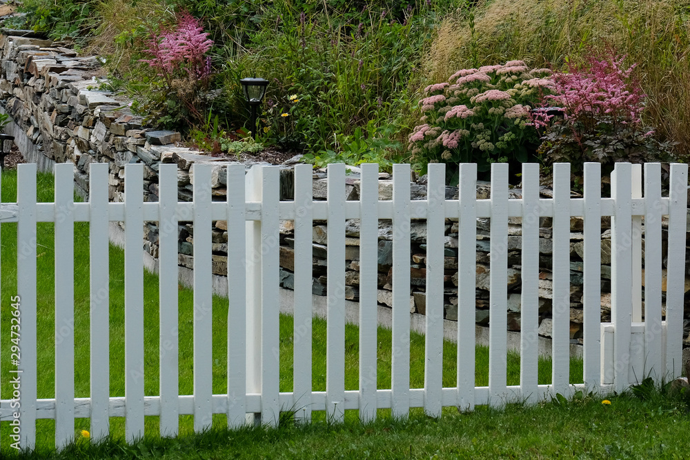 A country wooden white picket fence and gate divides gardens. There's a rock wall with pink flowers and shrubs on one side and green grass on the other side. 