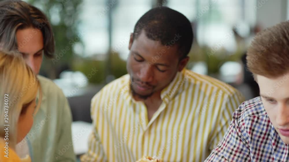 Camera panning left and right past diverse friends eating burger and working on homework assignment together while sitting in cafeteria together Camera panning left and right past diverse friends eating burger and working on homework assignment together while sitting in cafeteria together