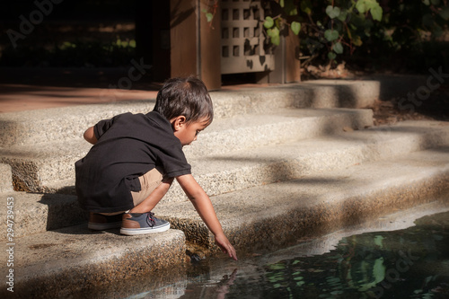 A young kid that is squatting down to reach the water from a pond and playing with the water during sunset in autumn.