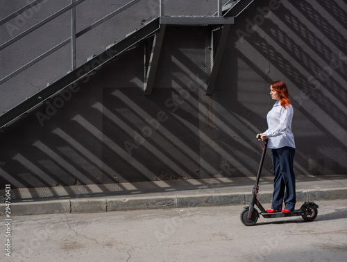 A red-haired girl in a white shirt drives an electric scooter along the wall. A business woman in a trouser suit and red high heels rides around the city in a modern car. Business woman on a scooter. © Михаил Решетников