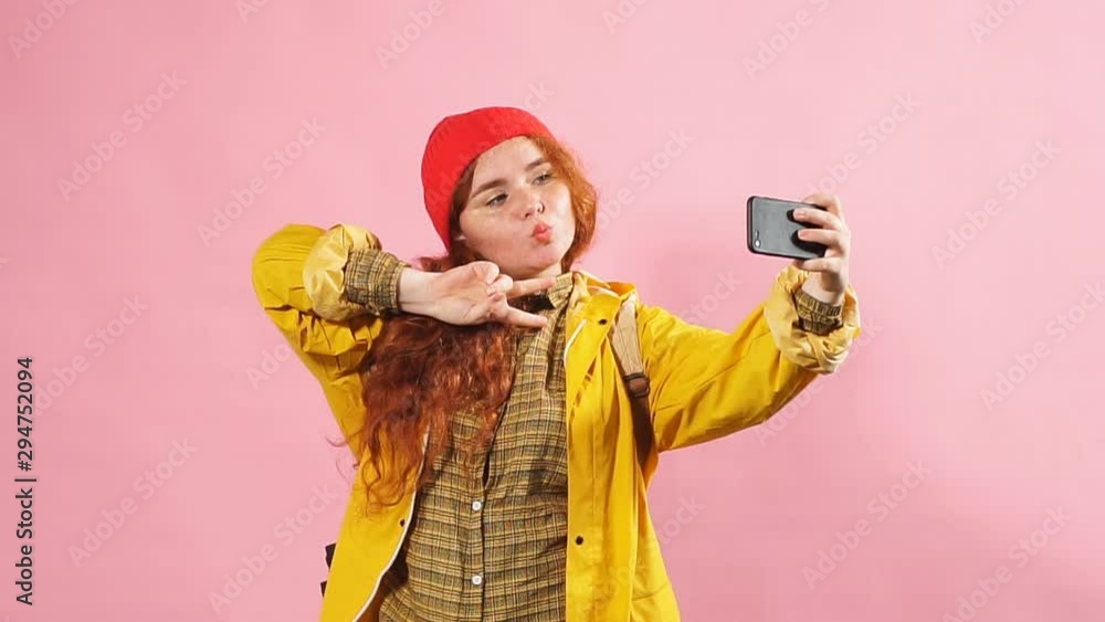 Young charming red-haired girl takes pictures of herself on the phone. Studio shot isolated on pink Young charming red-haired girl takes pictures of herself on the phone. Studio shot isolated on pink