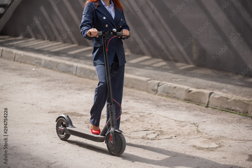 Obraz premium A red-haired girl in a jacket and trousers drives an electric scooter along the wall. A business woman in a pantsuit and red high heels rides around the city in a modern car. Office worker. Dress code