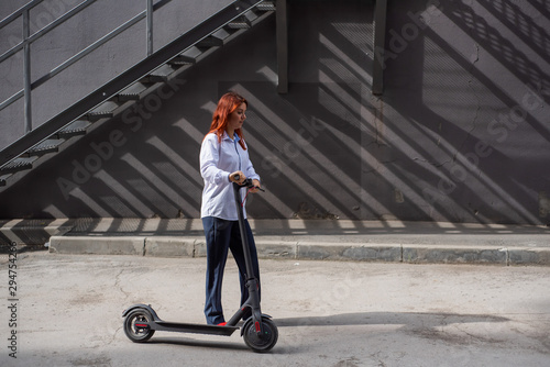 A red-haired girl in a white shirt drives an electric scooter along the wall. A business woman in a trouser suit and red high heels rides around the city in a modern car. Business woman on a scooter.