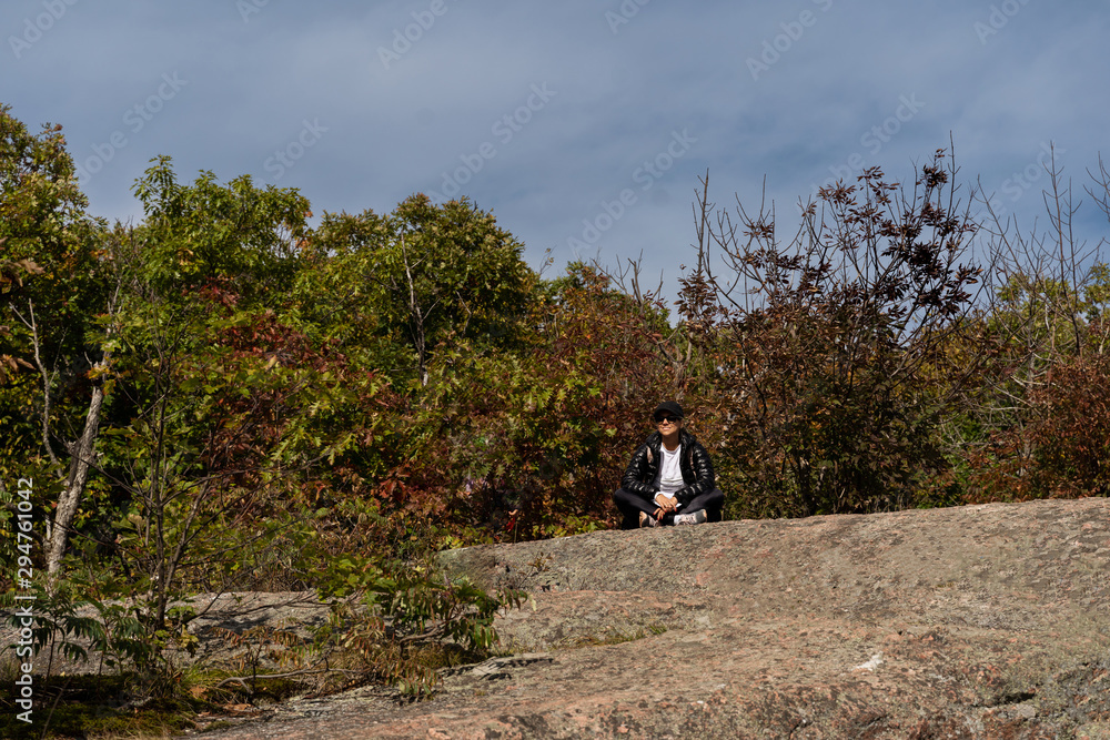 attractive blond woman dressed in black hiking jacket contemplating the view on top of rock in Gatineau Park, Quebec, Canada. Trees around her are turning red and orange in the fall. attractive blond woman dressed in black hiking jacket contemplating the view on top of rock in Gatineau Park, Quebec, Canada. Trees around her are turning red and orange in the fall.