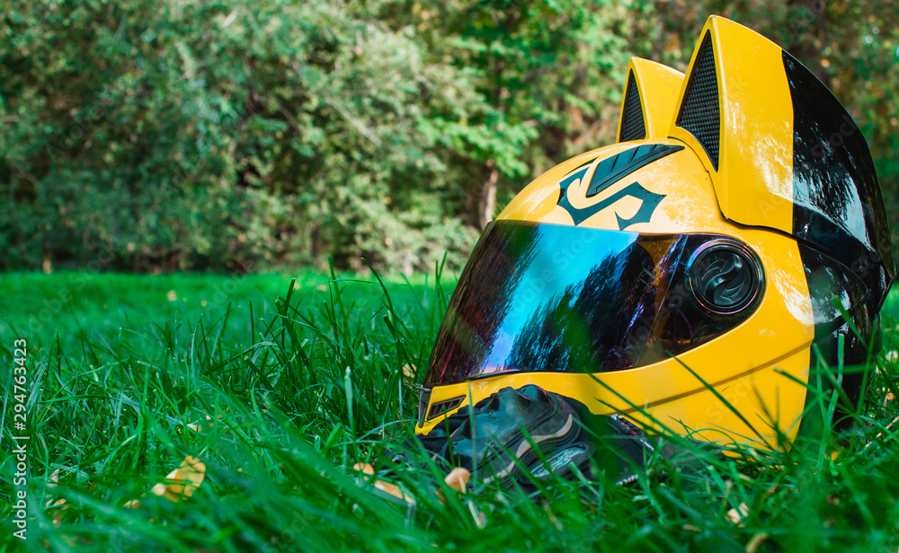 A yellow biker girl helmet from a motorcycle with mirror glass and ears and  black biker gloves lies on green grass against the background of a gray  motorcycle in autumn. Stock Photo