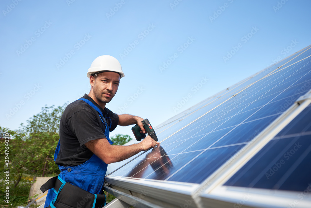 Technician installing solar panel to metal platform using electrical screwdriver on blue sky copy space background. Stand-alone solar system installation, efficiency and professionalism concept. Technician installing solar panel to metal platform using electrical screwdriver on blue sky copy space background. Stand-alone solar system installation, efficiency and professionalism concept.
