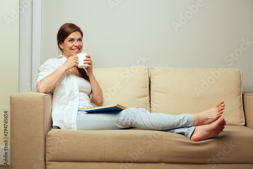 Smiling woman drinking coffee sitting on sofa with book.