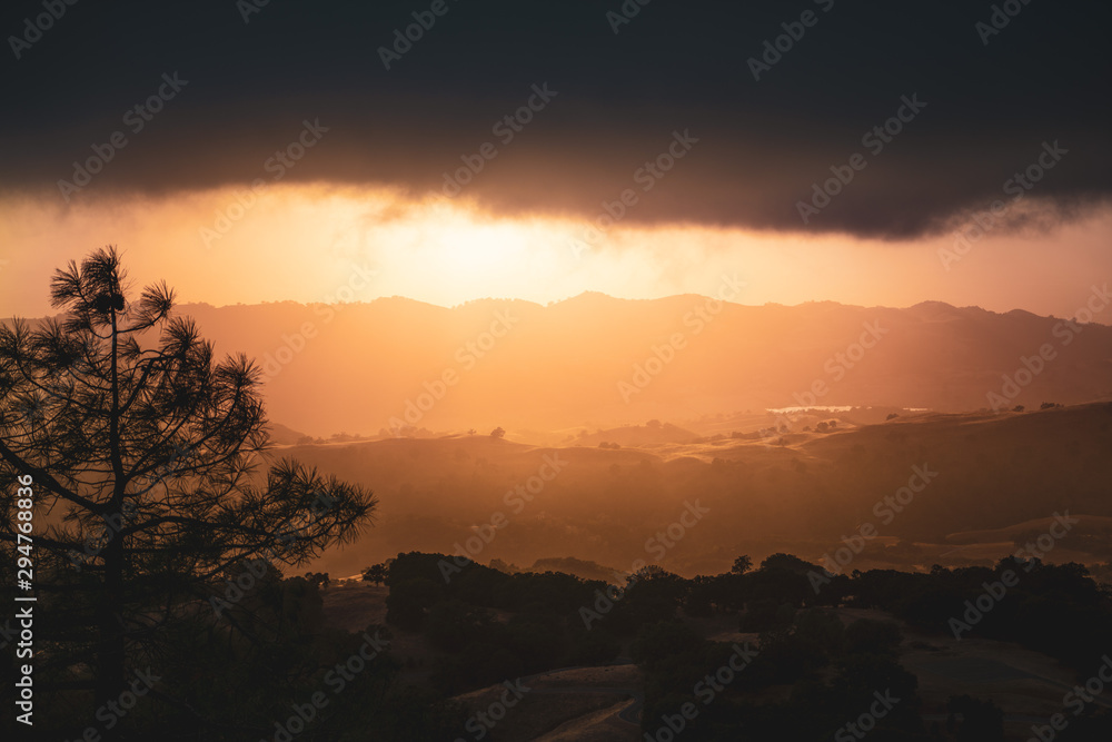 Dark and menacing storm clouds covering the sky at sunset; bright orange light illuminating hills and valleys in South San Francisco Bay Area; San Jose, California