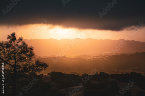 Dark and menacing storm clouds covering the sky at sunset; bright orange light illuminating hills and valleys in South San Francisco Bay Area; San Jose, California
