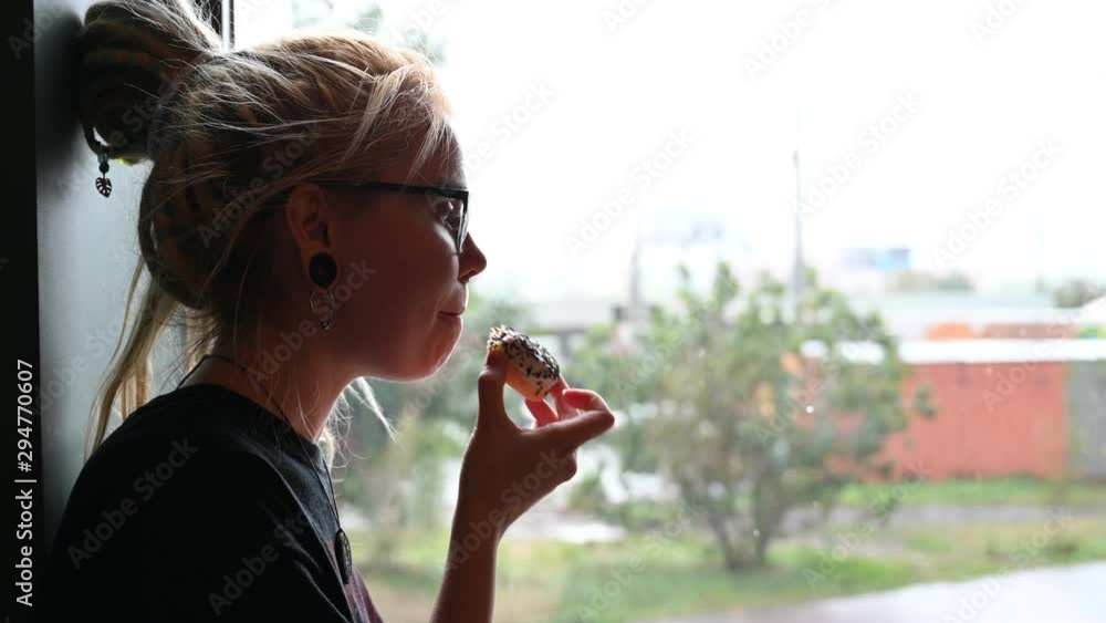 An unusual girl with dreadlocks on her head and tunnels in her ears drinks coffee and eats a donut in a cafe. A young woman in glasses is having breakfast while sitting by the window during the rain. An unusual girl with dreadlocks on her head and tunnels in her ears drinks coffee and eats a donut in a cafe. A young woman in glasses is having breakfast while sitting by the window during the rain.