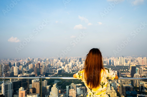 Woman wear yellow dress, Asian traveler standing with their backs turned and looking aside on balcony with beautiful modern big city view on background.