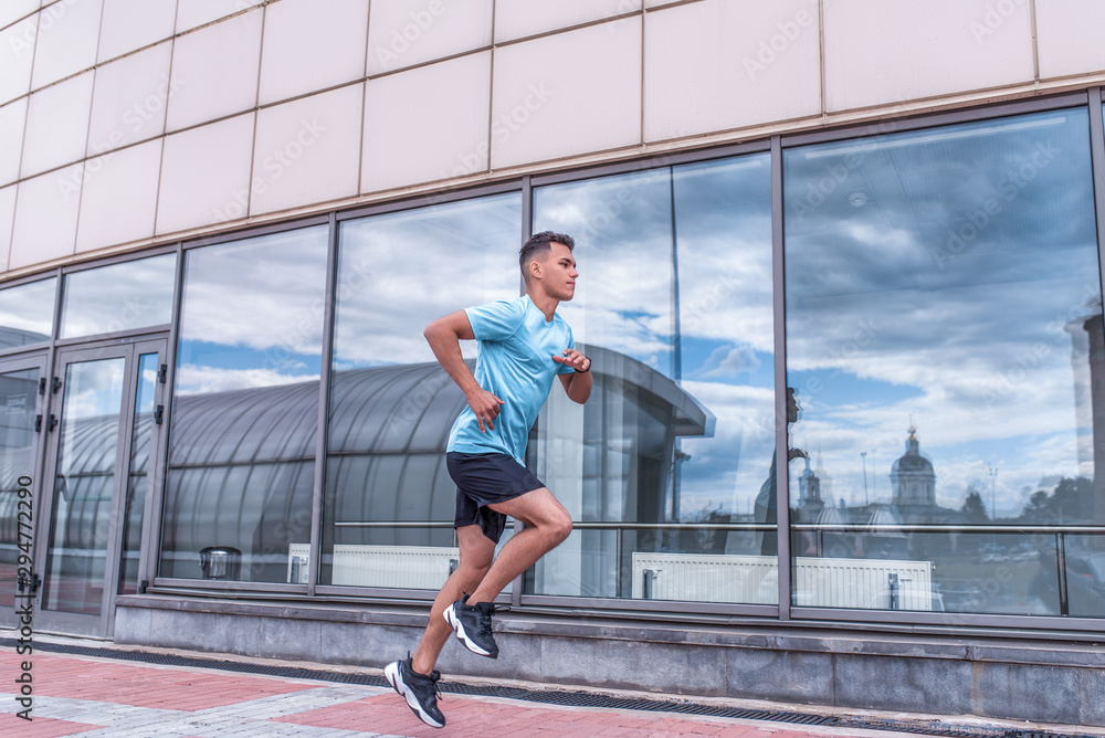 Obraz premium male athlete runs in morning, summer, spring in city, training in jump. Active lifestyle, workout, fitness in fresh air. Motivation for sports. Glass windows background. Free space.