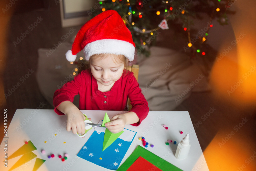 a little girl makes a card application of colored paper on the background of the Christmas tree. the concept of new year kids creativity a little girl makes a card application of colored paper on the background of the Christmas tree. the concept of new year kids creativity