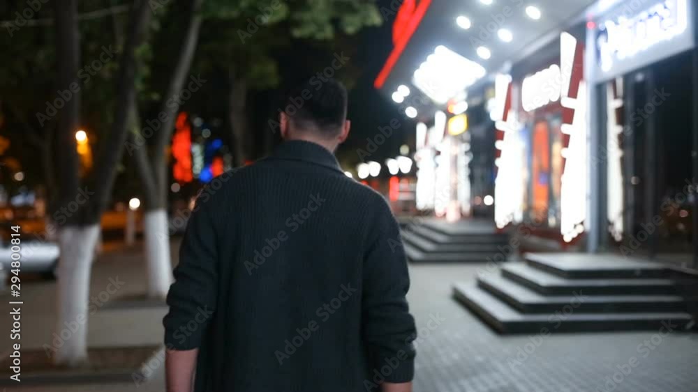 Back view of a handsome bearded man walking along the street at night, looking around , turning his head and waving hand. Slide camera Back view of a handsome bearded man walking along the street at night, looking around , turning his head and waving hand. Slide camera