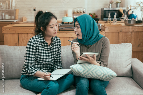 young asian girl team work partners discussing and making notes on sofa at college campus. muslim and chinese students studying together sitting on couch in dormitory. female roommates talking. photo