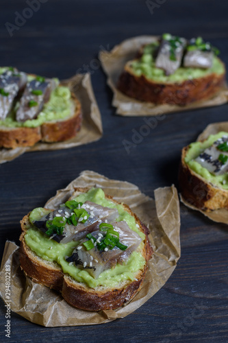 Avocado and herring sandwiches sprinkled with spring onions on grain bread top view on a paper napkin on a dark wooden background