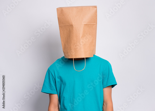 Portrait of teen boy with paper bag over head. Teenager cover head with bag posing in studio. Child pulling bag over head on gray background. photo