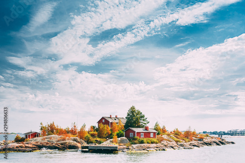 Suomi Or Finland. Beautiful Red Finnish Wooden Log Cabin House On Rocky Island Coast In Summer Sunny Evening. Lake Or River Landscape. Tiny Rocky Island Near Helsinki, Finland