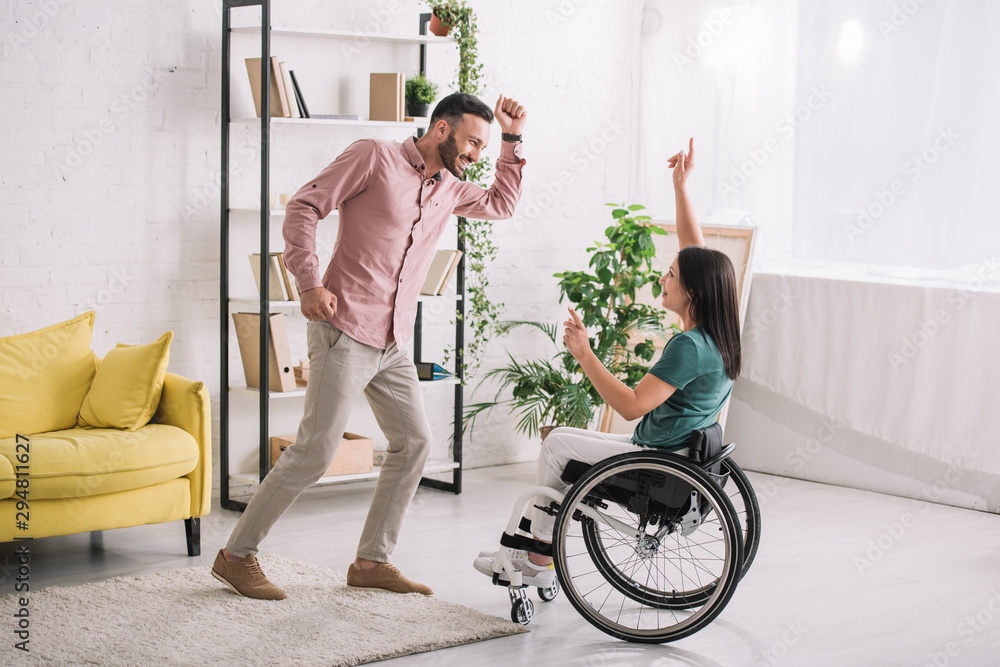 happy disabled woman on wheelchair dancing with cheerful boyfriend at home - obrazy, fototapety, plakaty 