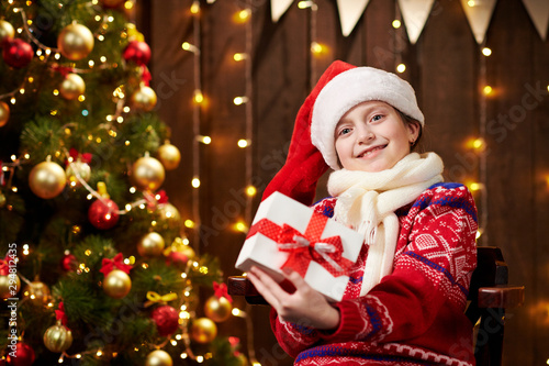 Cheerful santa helper girl with gift box sitting indoor near decorated xmas tree with lights, dressed in red sweater - Merry Christmas and Happy Holidays!