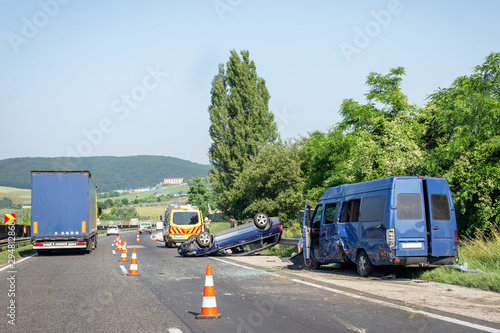 Car crash accident on highway. Damaged blue minibus after collision, overturned car and ambulance car on roadside. Traffic cones at accident site