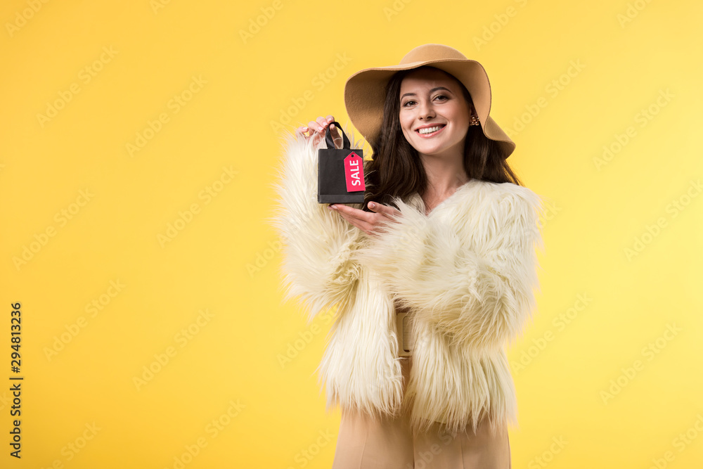 smiling elegant girl in faux fur jacket and hat holding shopping bag with sale lettering isolated on yellow smiling elegant girl in faux fur jacket and hat holding shopping bag with sale lettering isolated on yellow