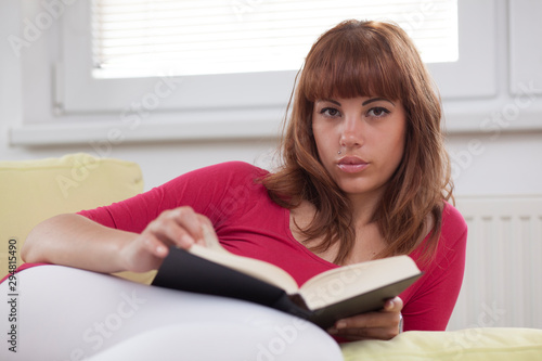 Brunette girl reading a black book seated on a sofa with window behind her, looking at the camera