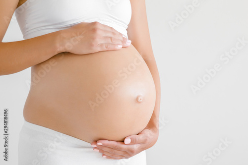Woman standing and touching with hands her naked big belly. Isolated on gray background. Emotional loving pregnancy time - 37 weeks. Baby expectation. Love, happiness and safety concept. Closeup. photo