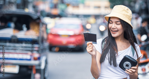 Traveller young Asian backpack woman wearing hat walking at Khao San road hands holding instant camera and film to take a picture, famous travelling landmark in Bangkok city of Thailand. 