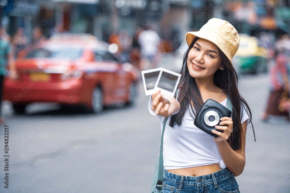Traveller young Asian backpack woman wearing hat walking at Khao San road hands holding instant camera and film to take a picture, famous travelling landmark in Bangkok city of Thailand. 