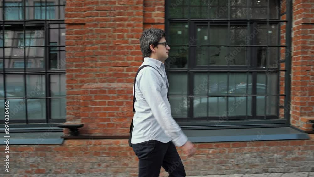 Middle gimbal profile shot. A serious young man walking fast from left to right along the street in eyeglasses wearing blue shirt on background of red brick building with windows
