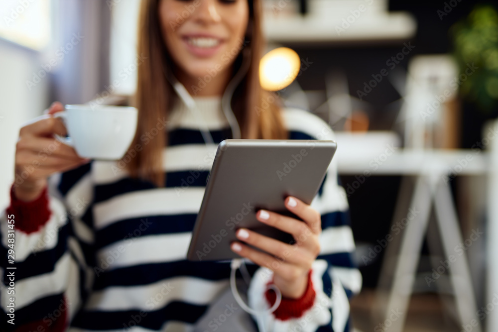 Beautiful smiling caucasian brunette dressed in sweater sitting on bed in bedroom, using tablet and drinking coffee in morning. Beautiful smiling caucasian brunette dressed in sweater sitting on bed in bedroom, using tablet and drinking coffee in morning.