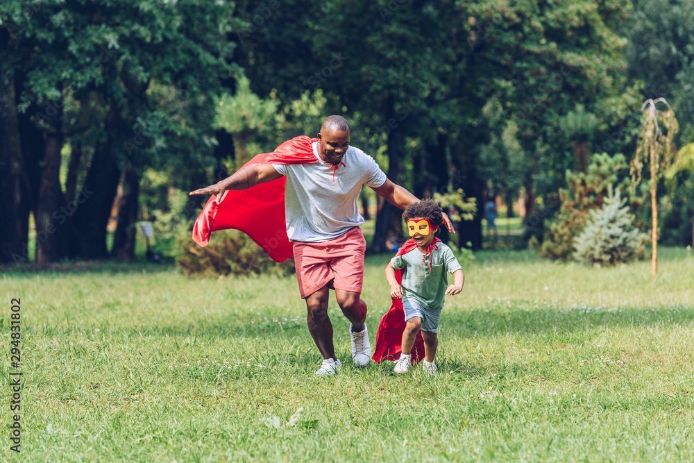 happy african american father and son running in costumes of superheroes in park - obrazy, fototapety, plakaty 