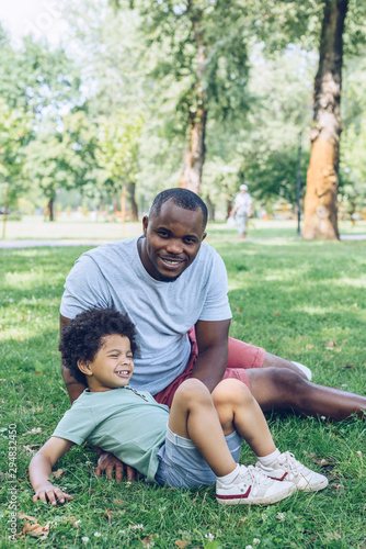 happy african american man smiling at camera while sitting on lawn near son in park
