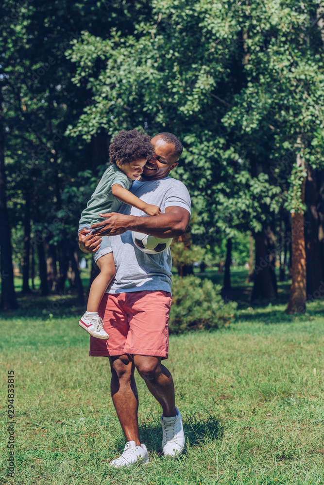 Fototapeta premium happy african american boy holding soccer ball while sitting on fathers hands