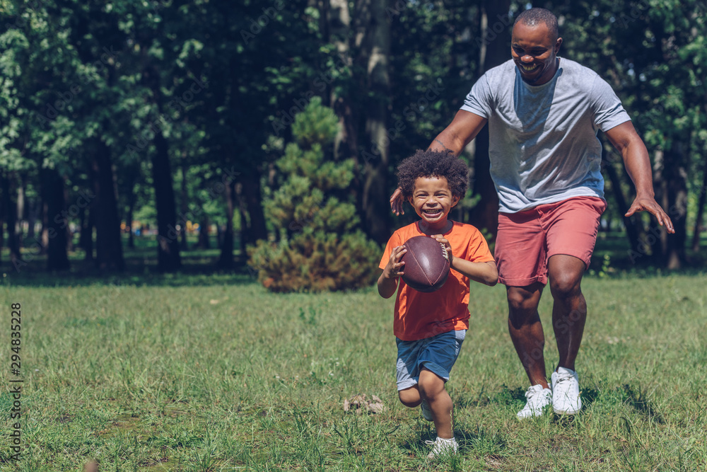 cute african american boy running with rugby ball while having fun with father in park