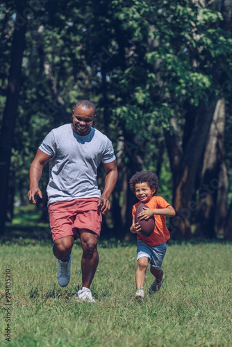 cheerful african american boy running with rugby ball while having fun with father in park