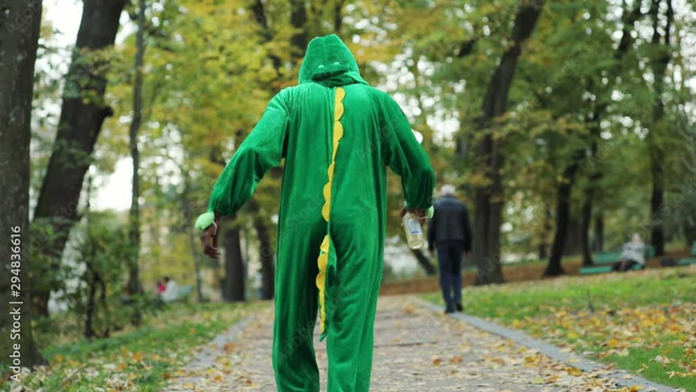 Back of green dinosaur male actorwalking slow in the park tottering carrying beer bottle. Drunken youth. Addictive generation. Back of green dinosaur male actorwalking slow in the park tottering carrying beer bottle. Drunken youth. Addictive generation.