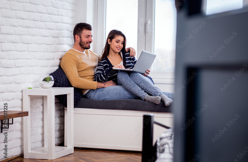 Obraz premium Young couple siting by the window and using at laptop spending time together at home.