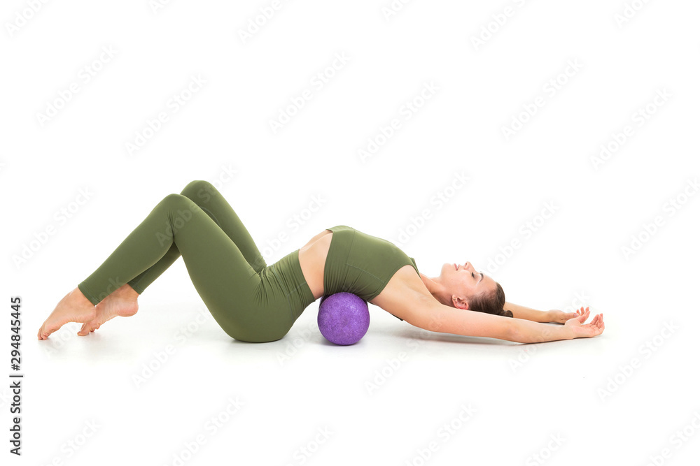 A beautiful young female gymnast with dark long hair stuffed into a bundle in a green sports elastic suit makes a stretch on various muscle groups with sporting inventory