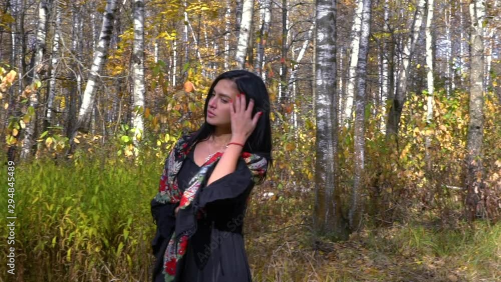A girl with black hair looks like a gypsy, she's all alone in the forest. Autumn yellow foliage around her. She is wearing a black dress and a mole on her lip. Cold autumn in Russia. A girl with black hair looks like a gypsy, she's all alone in the forest. Autumn yellow foliage around her. She is wearing a black dress and a mole on her lip. Cold autumn in Russia.