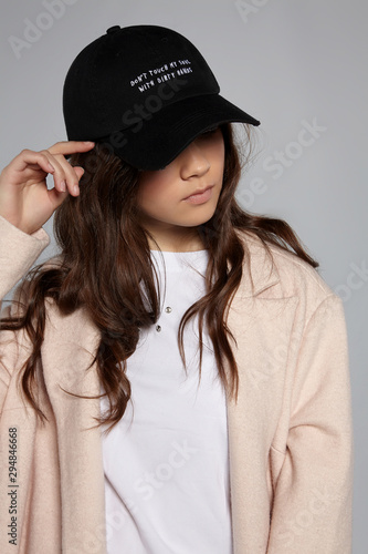 Medium close-up shot of young brown-haired lady in a white top with steel rivets, beige jacket and a black baseball cap with an embroidered lettering 